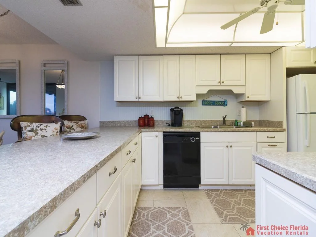White cabinetry and beige laminate countertops surround a black dishwasher and stainless sink beneath fluorescent ceiling lighting.