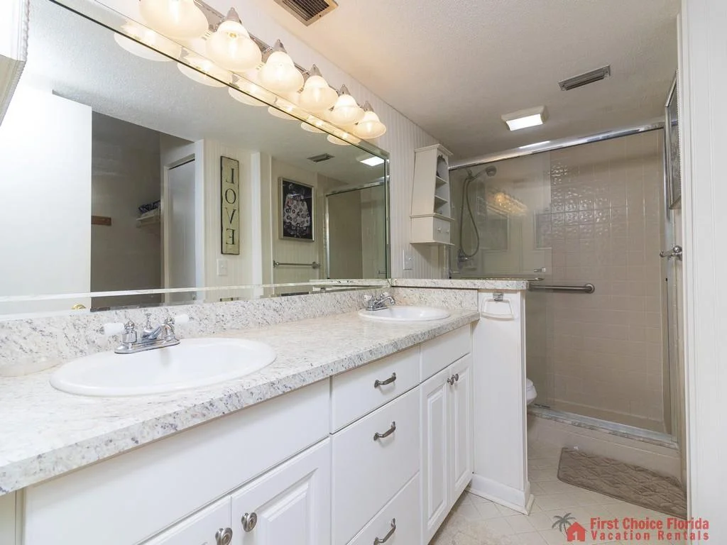 Dual sinks set in a speckled granite countertop with white cabinetry and a glass-enclosed tiled shower.