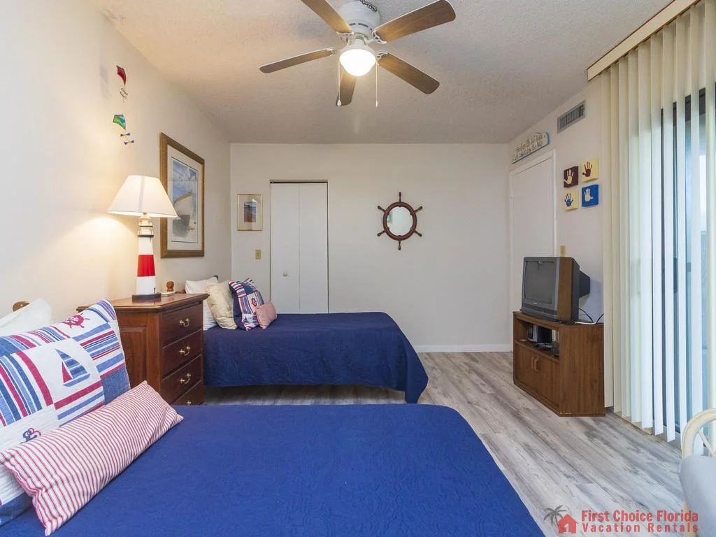 Two twin beds with navy quilted covers and nautical pillows sit opposite a wood TV cabinet and sliding vertical blinds.