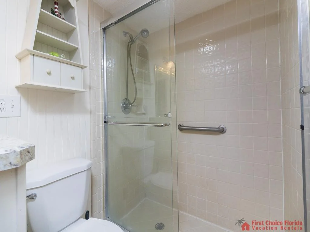 Bathroom features a glass-enclosed shower with beige tile walls, a chrome grab bar, and a white toilet below a small wall shelf.
