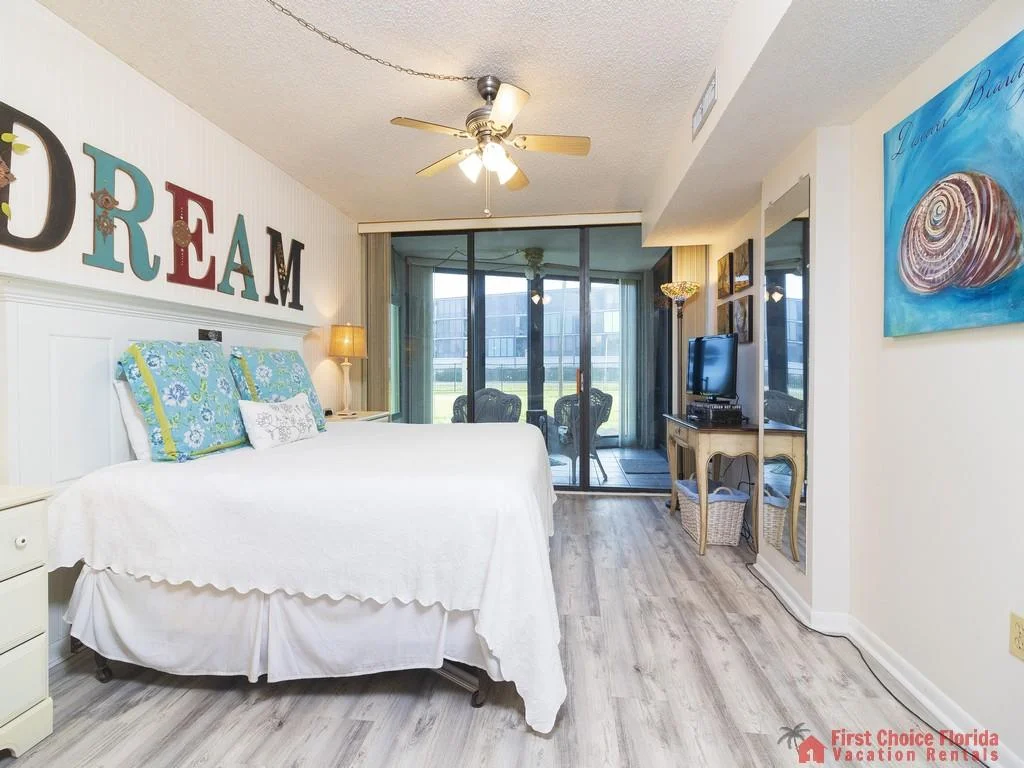 King bed with floral pillows faces sliding glass doors opening to a covered patio with black wicker chairs.