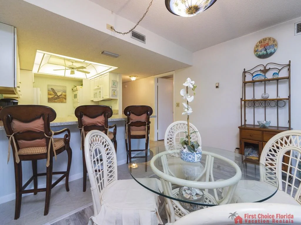 Three dark wooden barstools with cushioned seats line the kitchen counter opposite a glass dining table with white wicker chairs.
