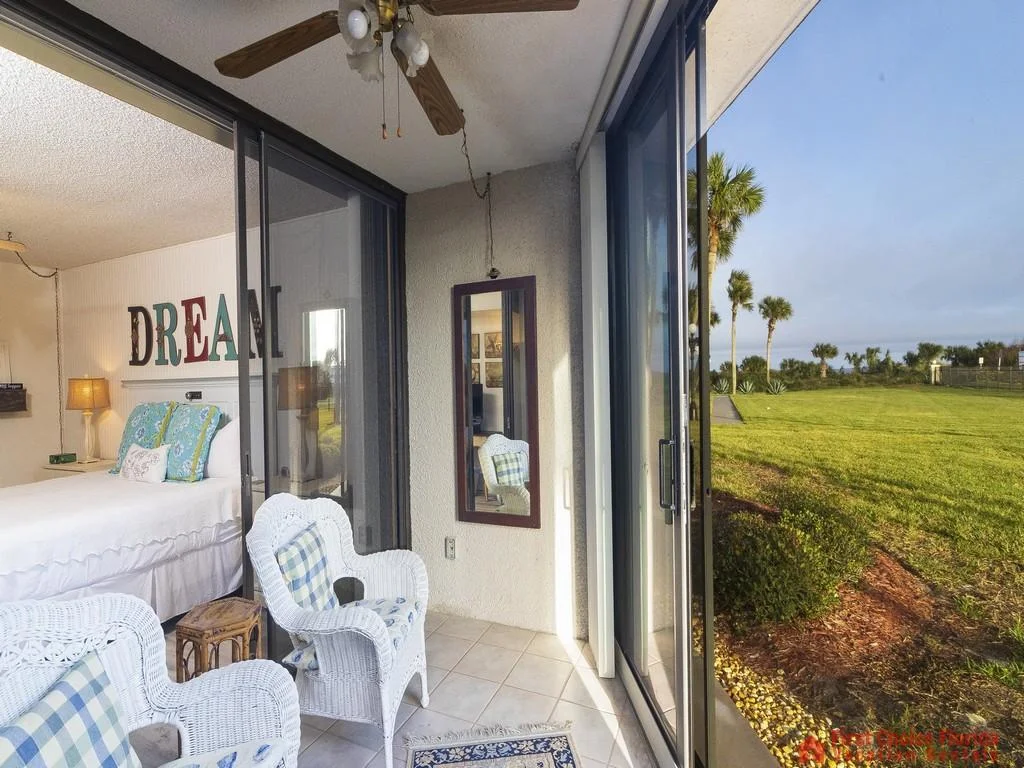 Sunroom with white wicker chairs and plaid cushions overlooks green lawn and palm trees through sliding glass doors.