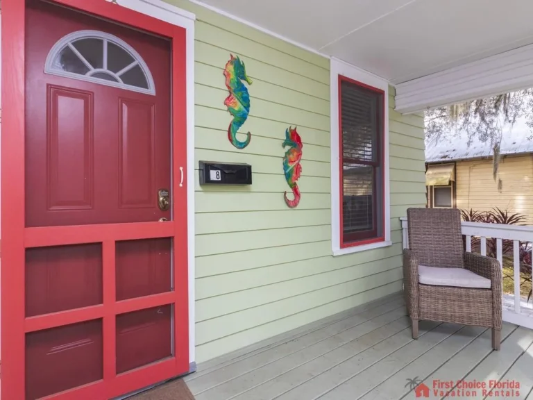 Red-framed door and window contrast with pale green siding, accented by colorful seahorse wall art and wicker chair.