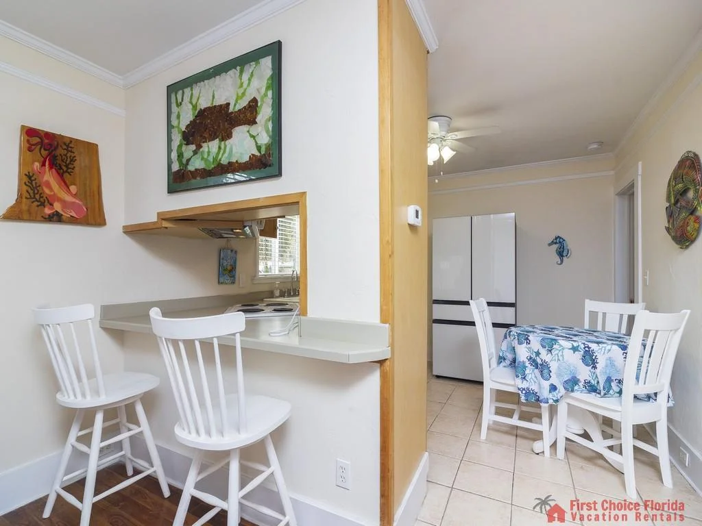 White bar stools line the kitchen pass-through overlooking a tiled dining area with floral tablecloth.