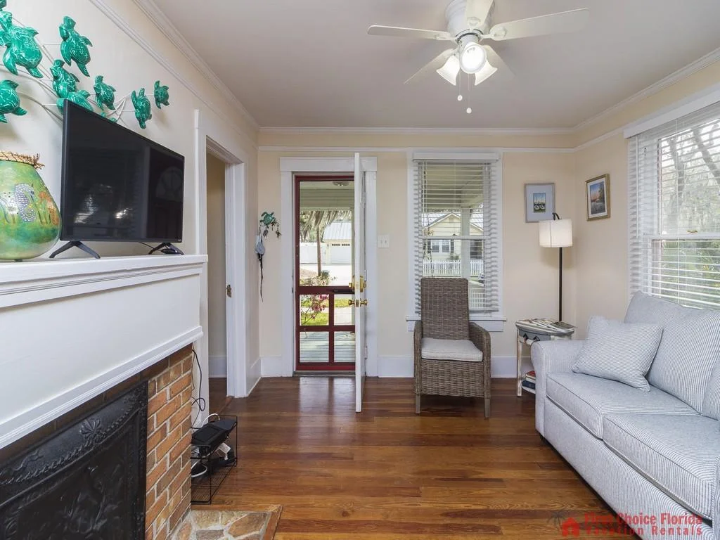 Light wood floors ground the living room with a striped gray sofa, wicker armchair, and white-trimmed windows.