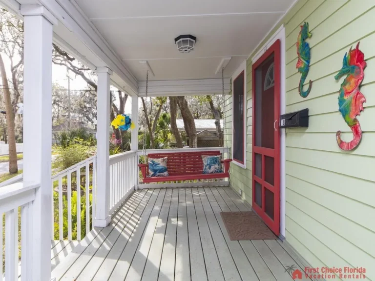 Wide front porch features pale gray wood planks, white railings, a red hanging bench, and colorful seahorse wall art.