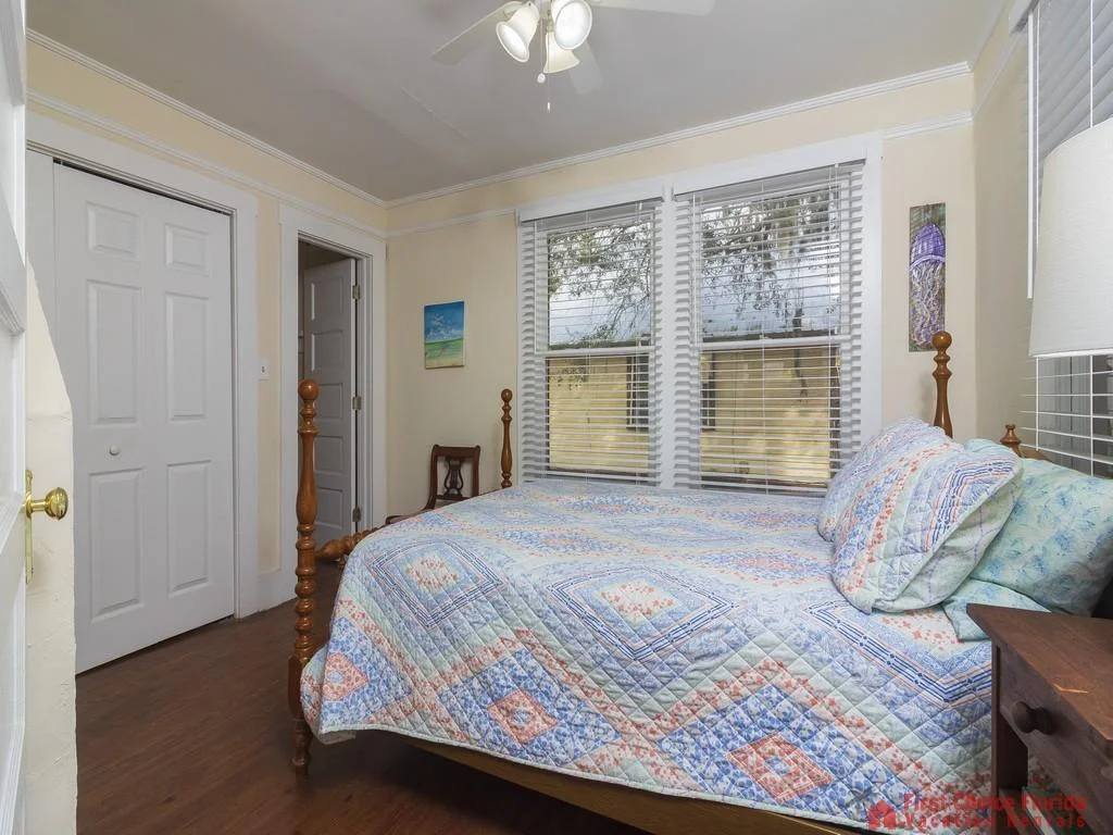 Four-poster bed with patterned quilt sits beside three large windows with white blinds and cream walls.