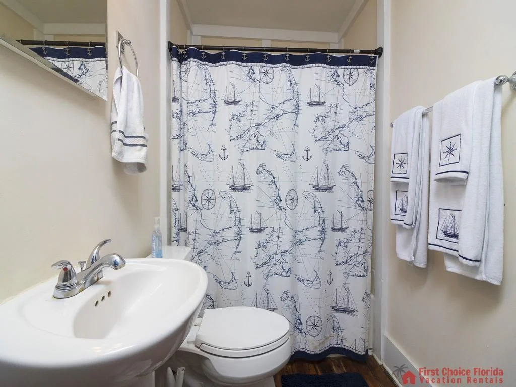 Bathroom with nautical-themed shower curtain, white sink, toilet, and embroidered white towels on wall rack.