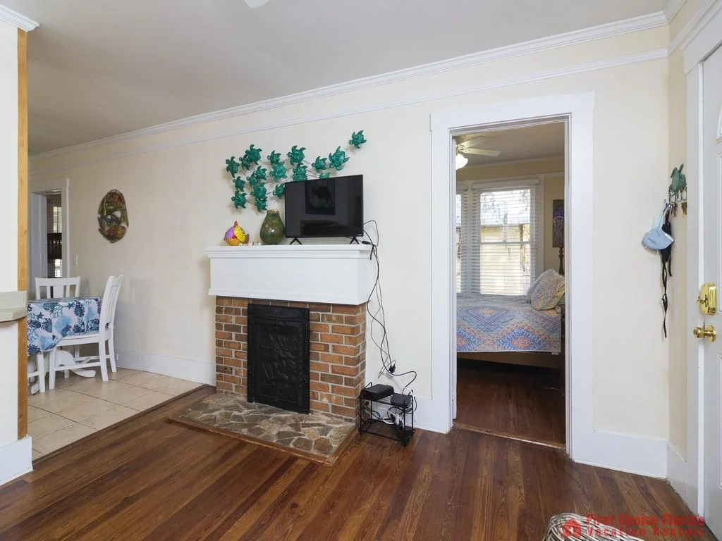 Brick fireplace with white mantel holds a TV, surrounded by hardwood floors and a doorway to the bedroom.