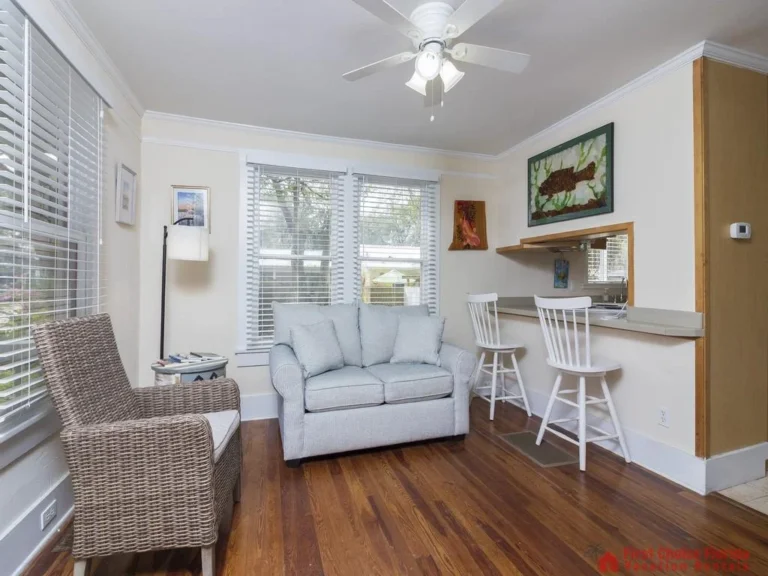 Wicker armchair and light blue loveseat face the kitchen pass-through with white bar stools and fish artwork.