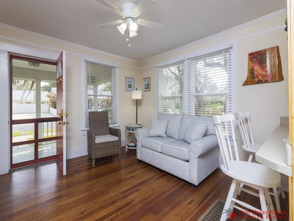 Natural light fills the living room with hardwood floors, a gray loveseat, wicker armchair, and white bar stools.