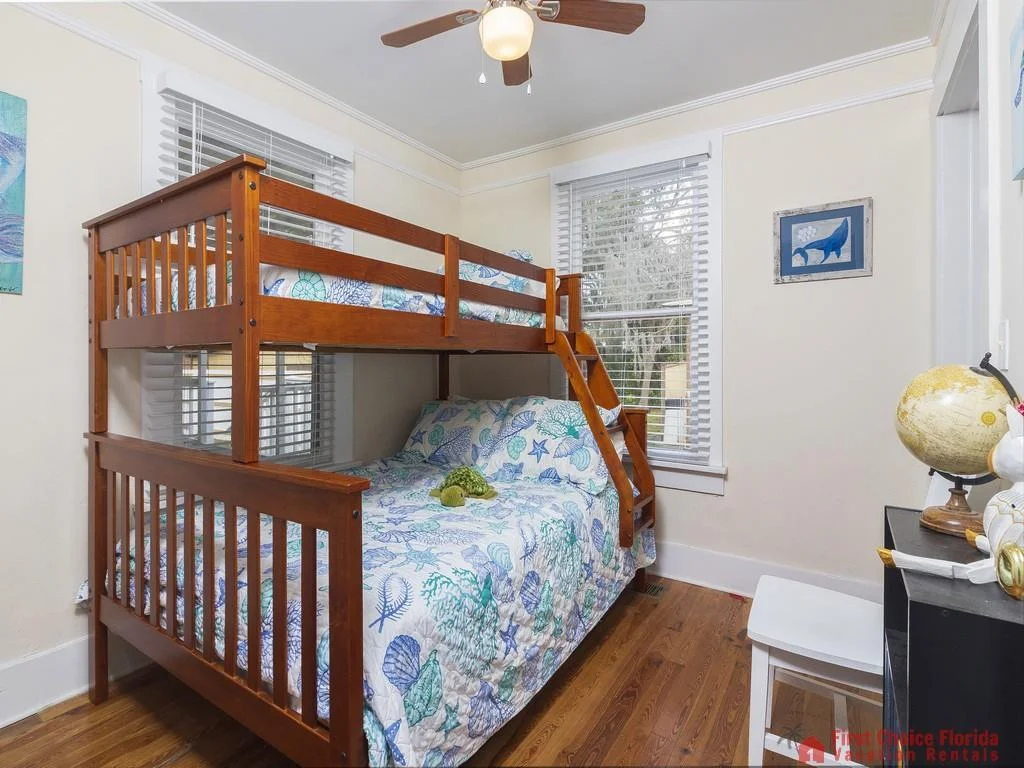 Wood bunk bed with seashell-patterned bedding sits beneath two windows with white blinds in the bedroom.