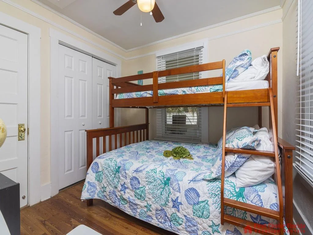 Wood-framed bunk bed with sea-themed quilt and pillows sits beneath a ceiling fan near two windows.