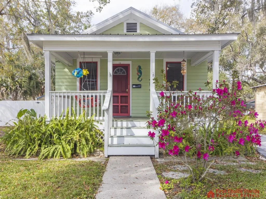Front exterior shows a pale green cottage with white trim, red door, porch swing, and blooming pink azalea bush.