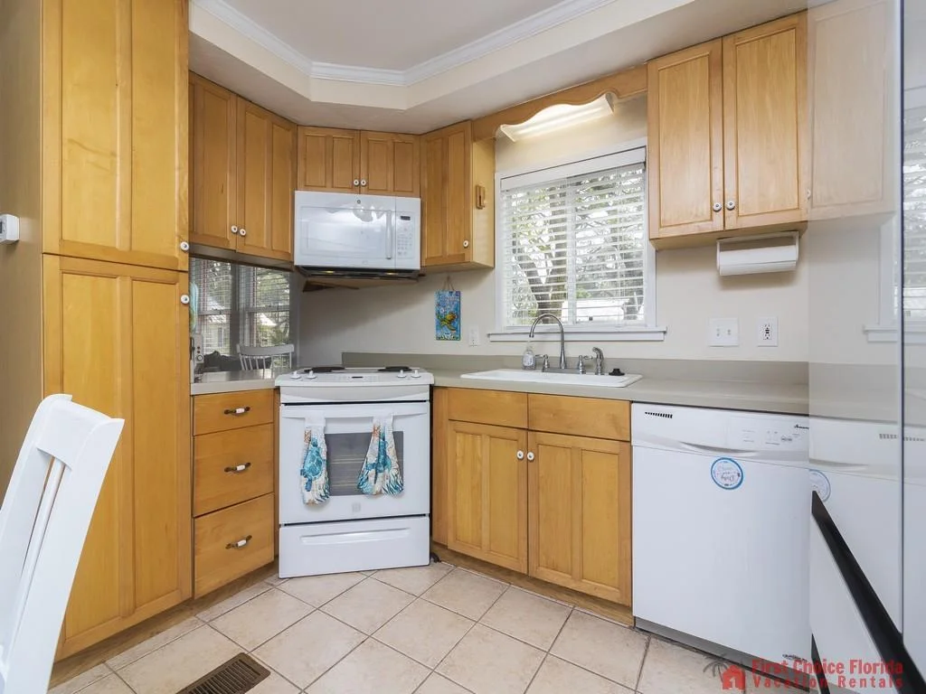 A window with white blinds overlooks the sink and light wood cabinetry, with a dishwasher and oven below.