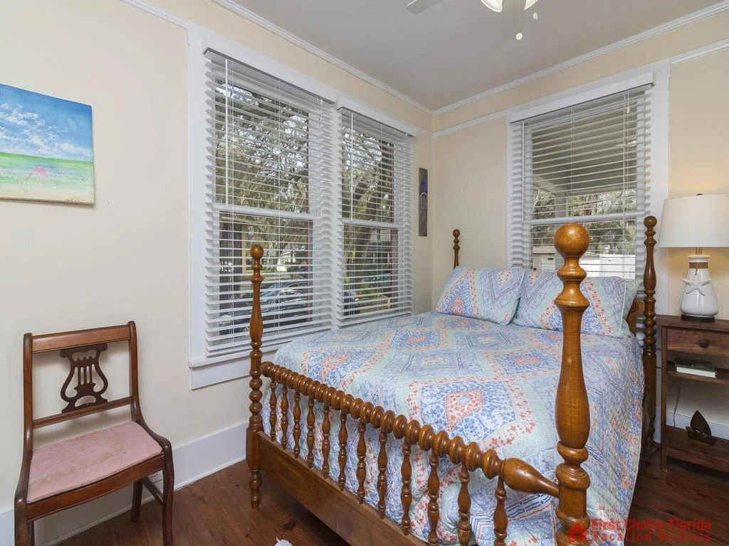 Turned wood bedposts frame a patterned quilted bedspread beneath windows with white blinds.