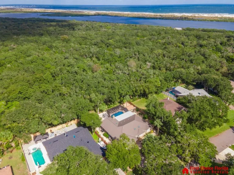Aerial view of the house surrounded by dense greenery, neighboring homes, and waterways stretching to the ocean horizon.