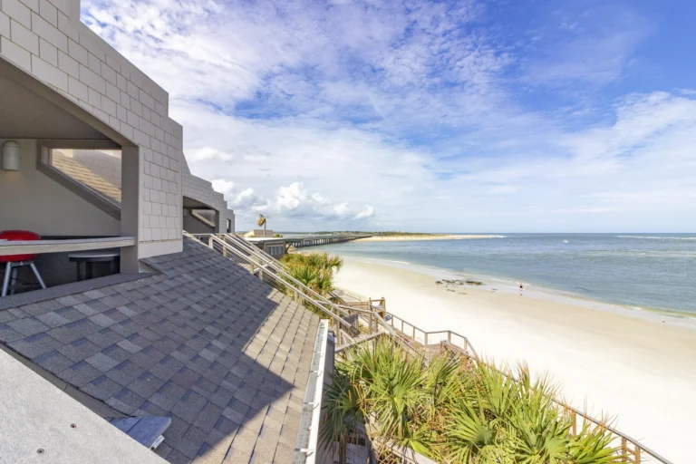 Oceanfront view from the penthouse balcony shows sandy beach, blue water, palm trees, and a distant bridge under a partly cloudy sky.