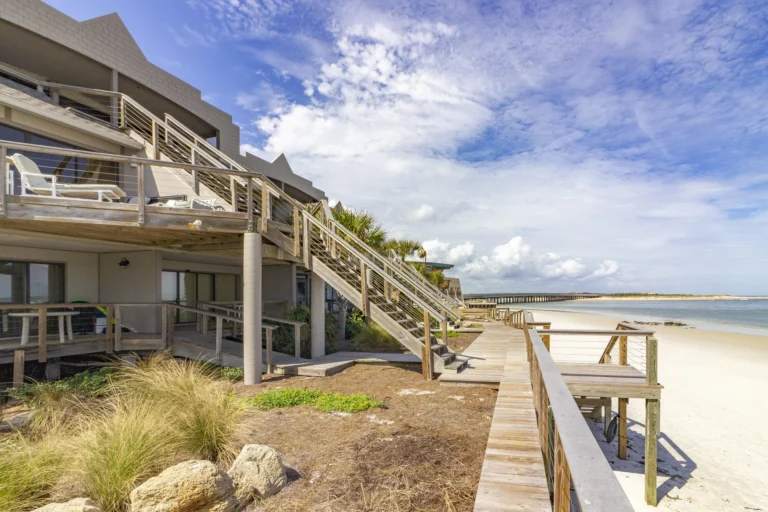 Wooden boardwalk and stairs lead up to oceanfront condos with sandy beach and calm blue water views.