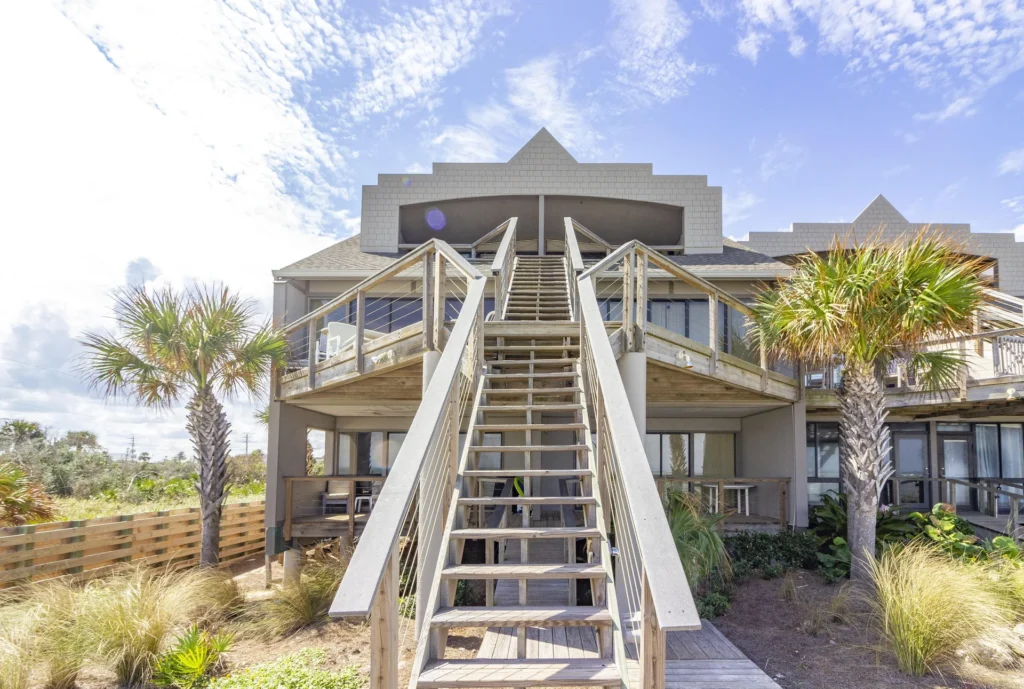 Exterior view of the two-story condo featuring a central wooden staircase, flanked by palm trees and sandy landscaping.