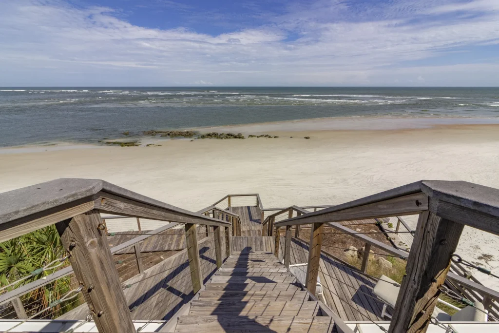 Sunlight casts shadows on the weathered wood steps and railing leading down to the sandy beach and ocean.