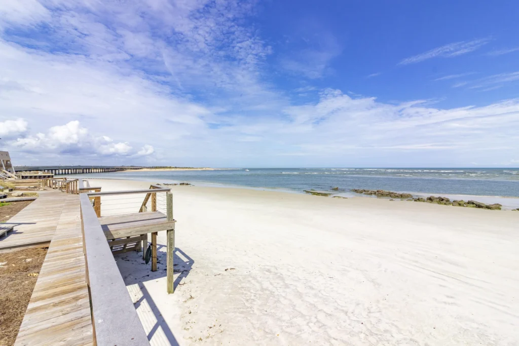 Wooden boardwalk with cable railing runs alongside white sandy beach and calm ocean under a partly cloudy sky.