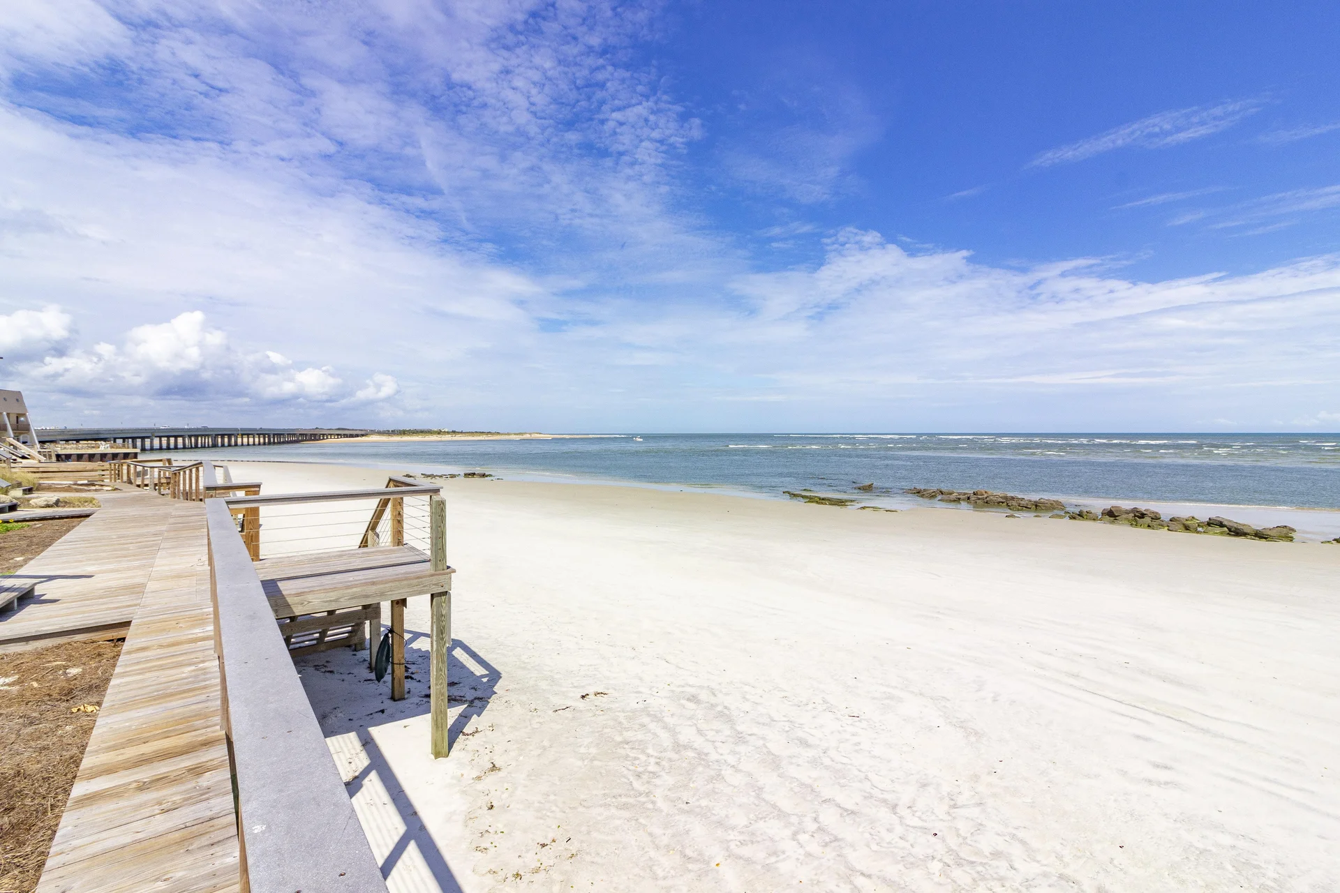 Wooden boardwalk with cable railing runs alongside white sandy beach and calm ocean under a partly cloudy sky.