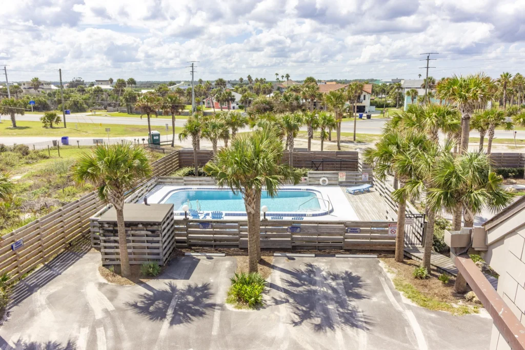 Gated outdoor pool area with white concrete deck, blue lounge chairs, surrounded by palm trees and wooden fencing.