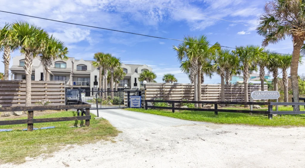 Gated entrance to the condo complex flanked by palm trees, wooden fencing, and a gravel driveway.