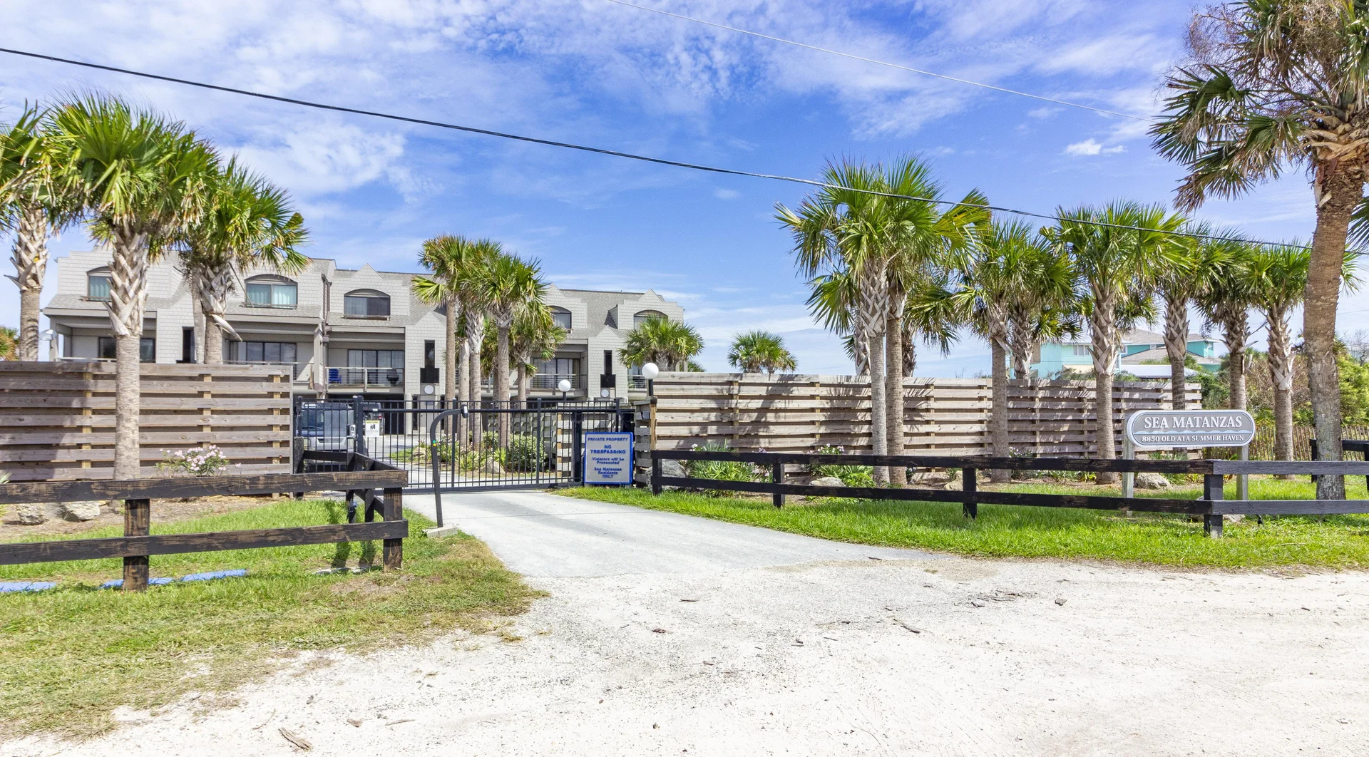 Gated entrance to the condo complex flanked by palm trees, wooden fencing, and a gravel driveway.