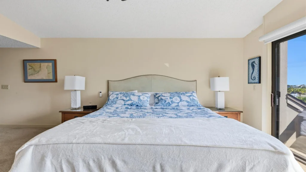 King bed with seashell-patterned blue quilt and white blanket between two wooden nightstands with white lamps.