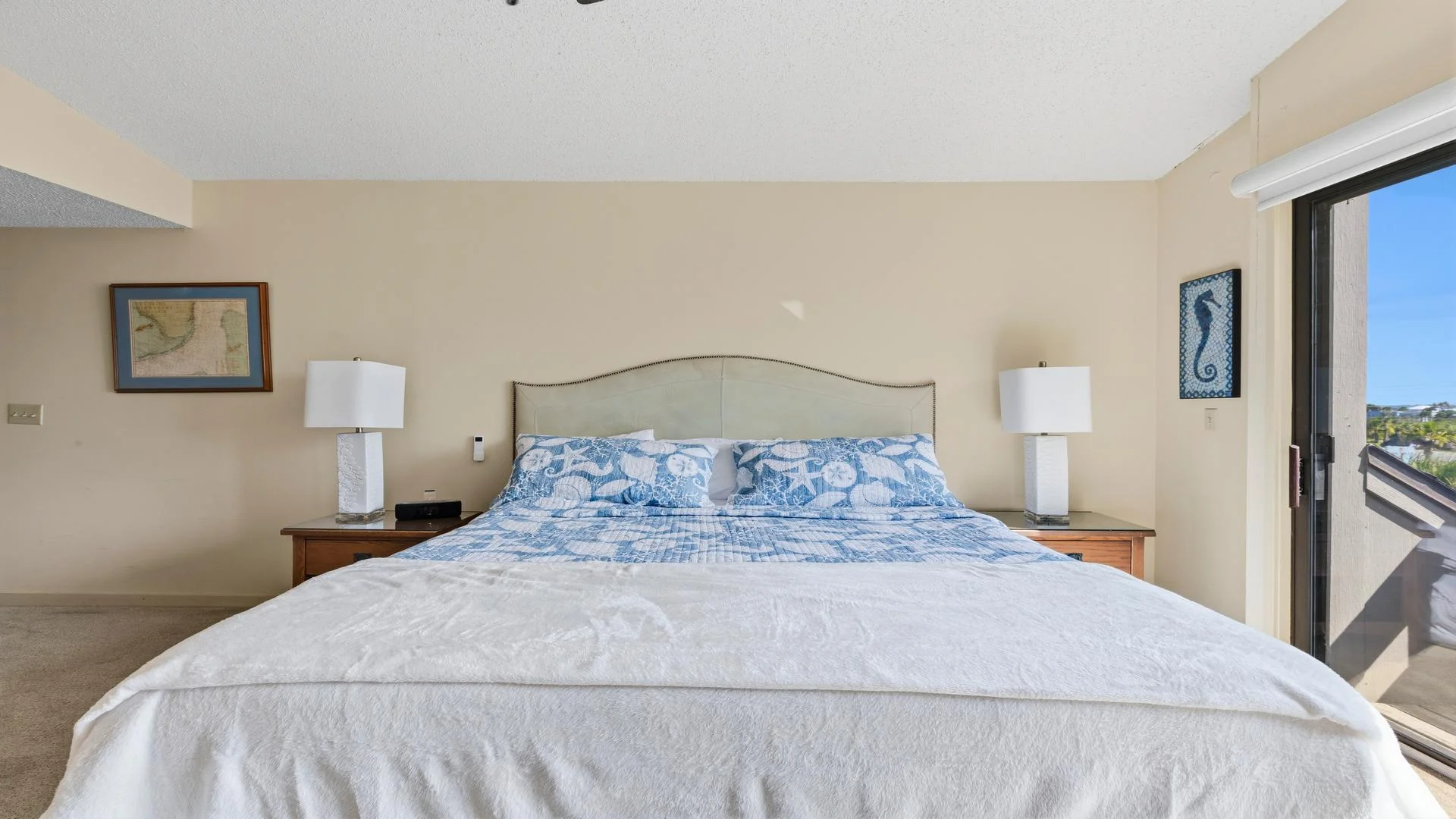King bed with seashell-patterned blue quilt and white blanket between two wooden nightstands with white lamps.