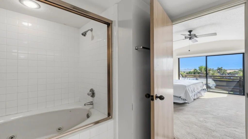 White tiled bathtub with sliding glass door next to a bedroom featuring wall-to-wall carpet and sliding glass doors.