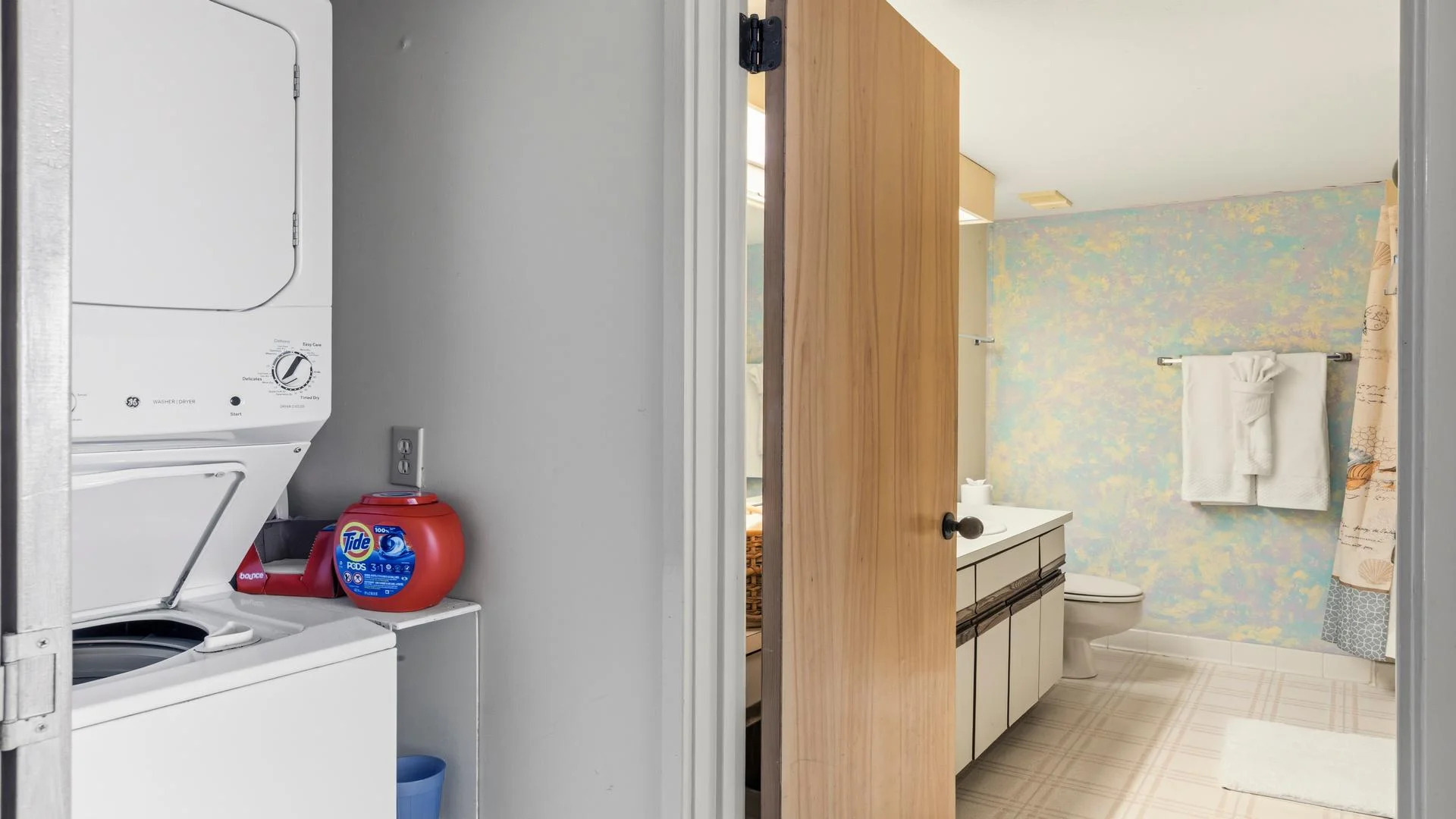 Stacked white washer and dryer sit in a compact laundry closet adjacent to a bathroom with pastel mottled walls.