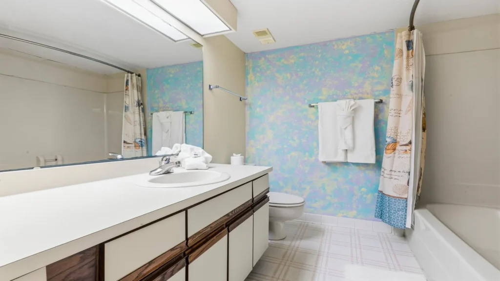 Bathroom with white laminate countertop, wood-trimmed vanity, pastel textured wall, and seashell-patterned shower curtain.