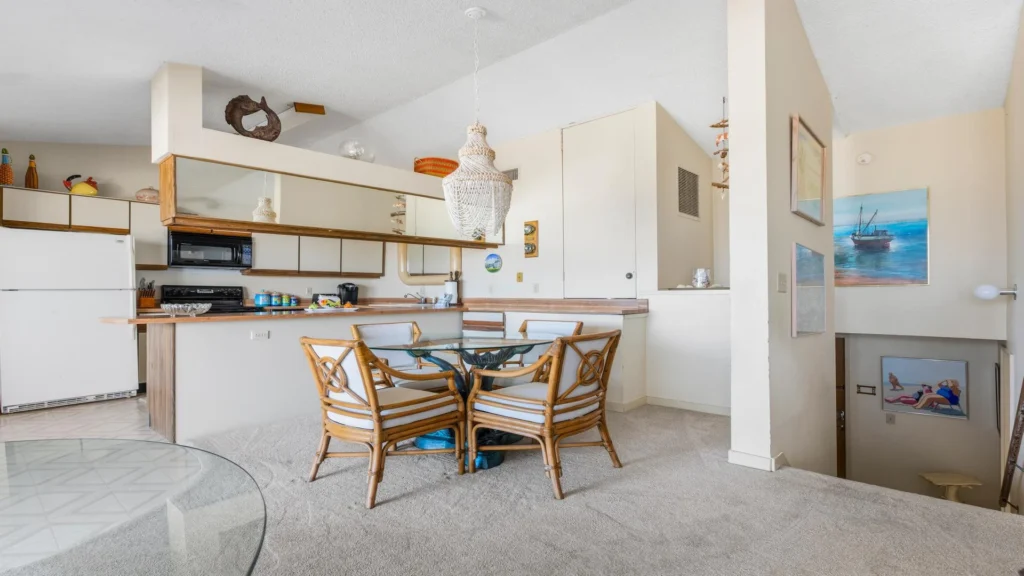 Glass-top dining table with four cushioned rattan chairs sits adjacent to a kitchen bar with wood trim and white appliances.
