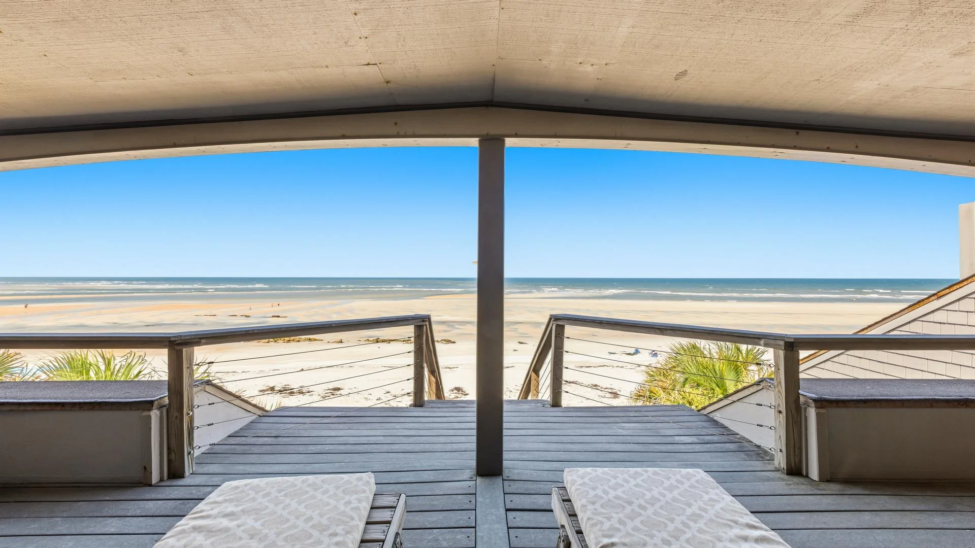Covered upper deck with wide gray wood planks, cable railing, and two lounge chairs facing the sandy beach and ocean.
