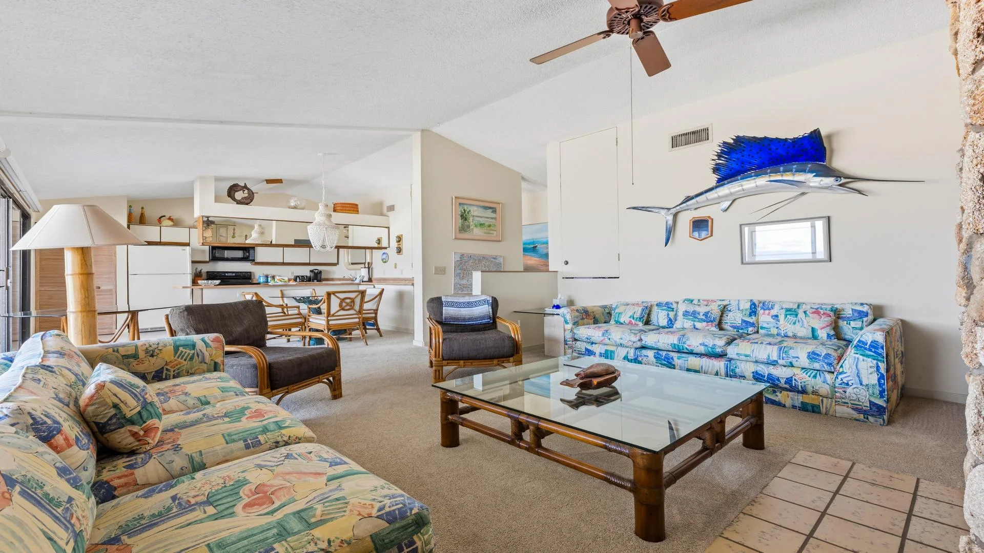 Living room features patterned sofas, bamboo armchairs, glass coffee table, and a mounted blue sailfish on cream walls.