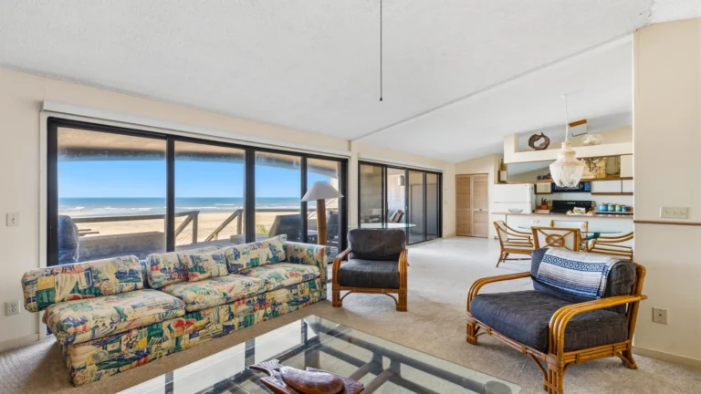 Living room features patterned sofa, rattan armchairs, glass coffee table, and ocean views through sliding windows.