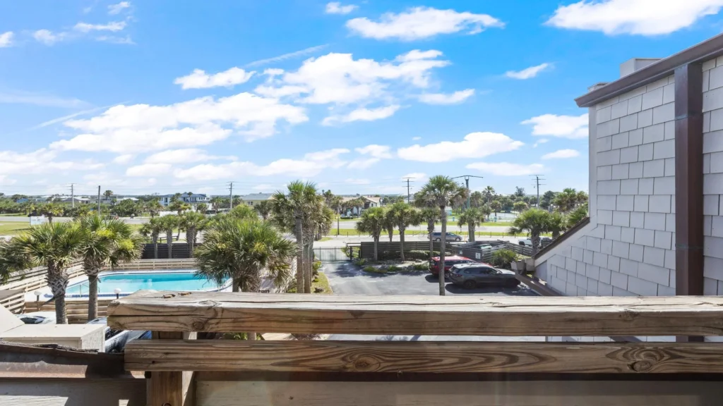 View over a wooden balcony railing reveals a palm tree-lined pool, parking area, and distant residential homes.