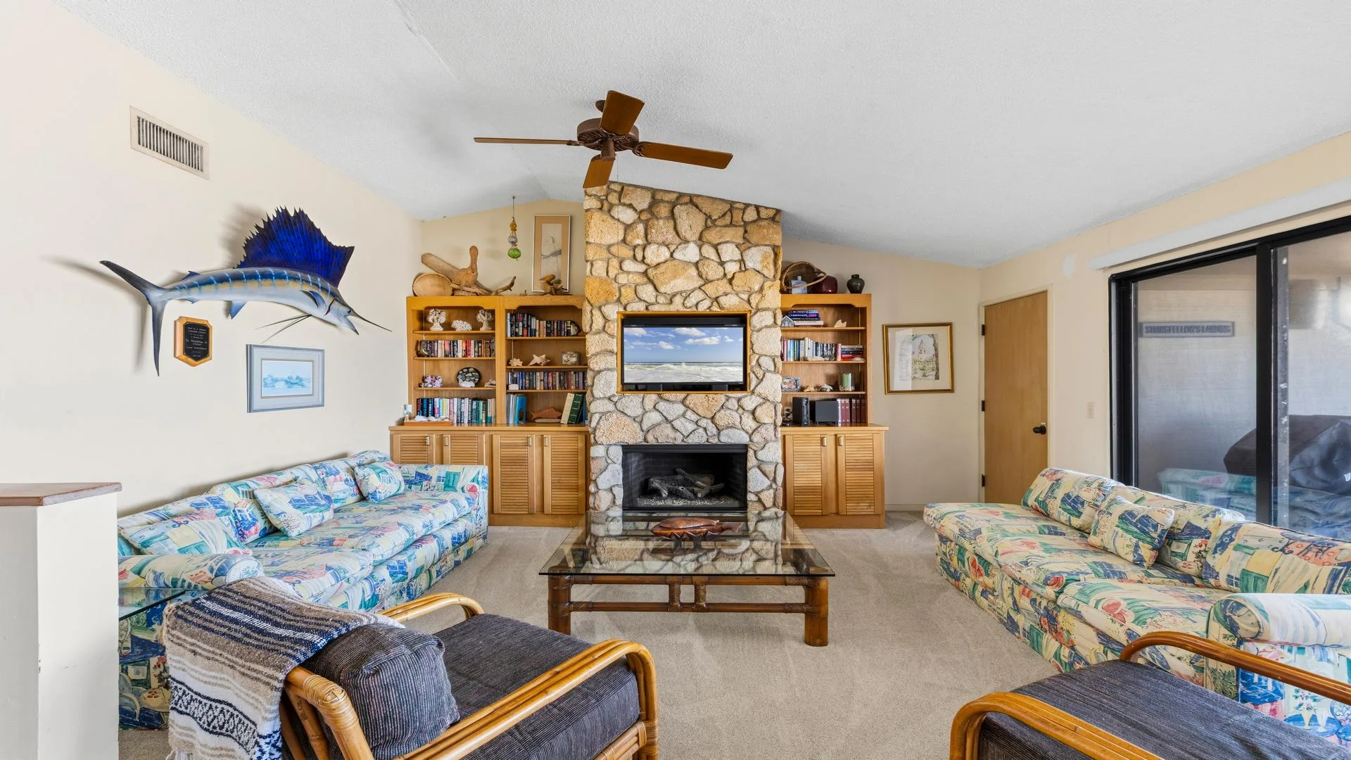 Stone fireplace with built-in wooden shelves flanks a glass coffee table between patterned sofas and rattan armchairs.