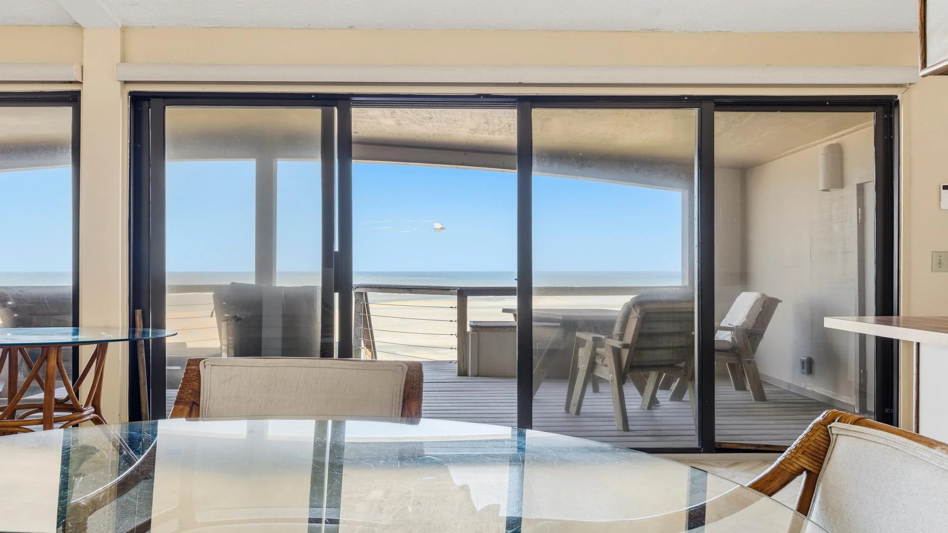 Glass sliding doors open to a covered deck with wooden Adirondack chairs facing the sandy beach and ocean.