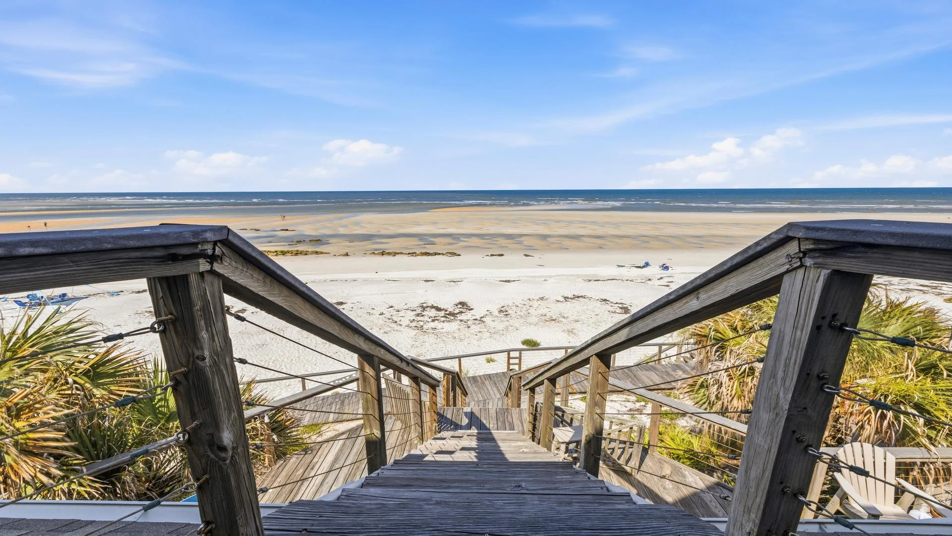 Wooden handrails with metal cables frame the descent to the beach, where sand meets shallow tidal pools.