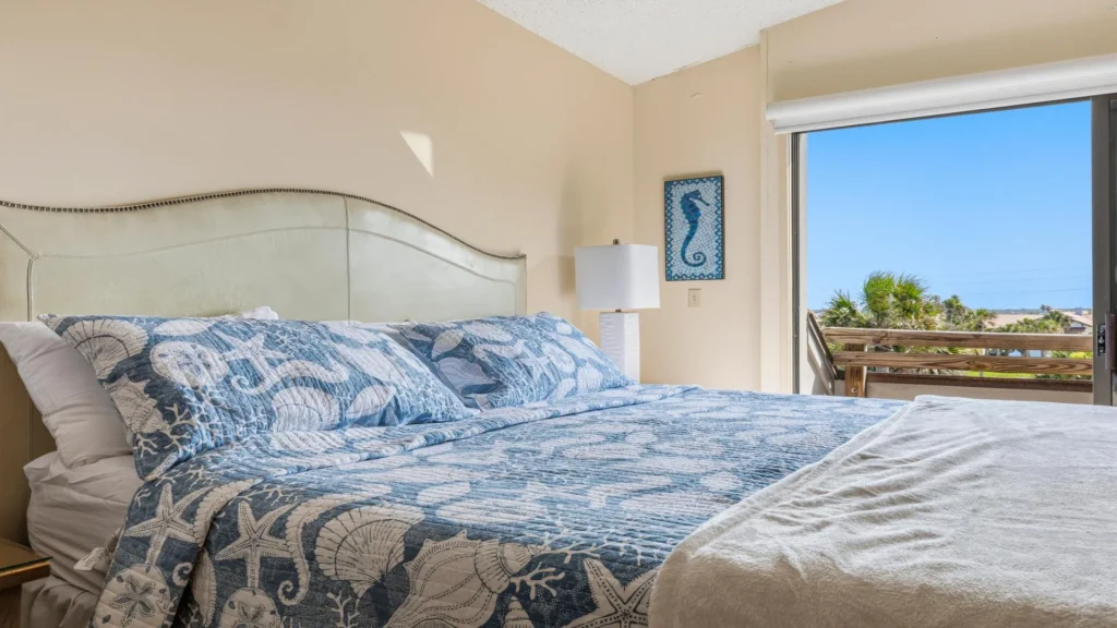 Light cream leather headboard with nailhead trim frames a king bed dressed in blue seashell quilt next to sliding glass doors.