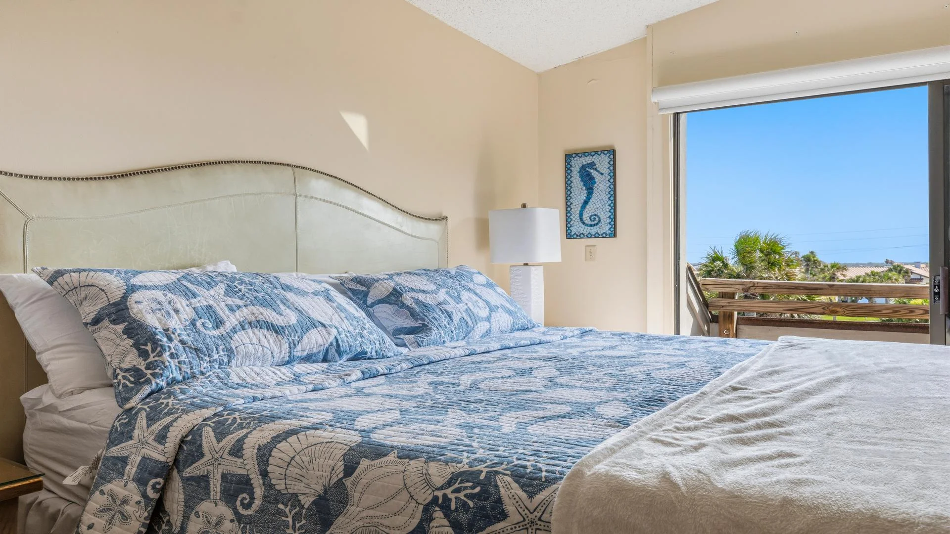 Light cream leather headboard with nailhead trim frames a king bed dressed in blue seashell quilt next to sliding glass doors.