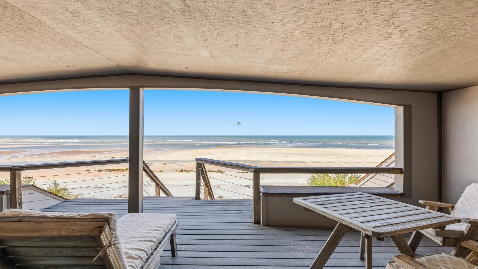 Shaded covered patio with wooden deck furniture overlooks sandy beach and calm ocean waves.
