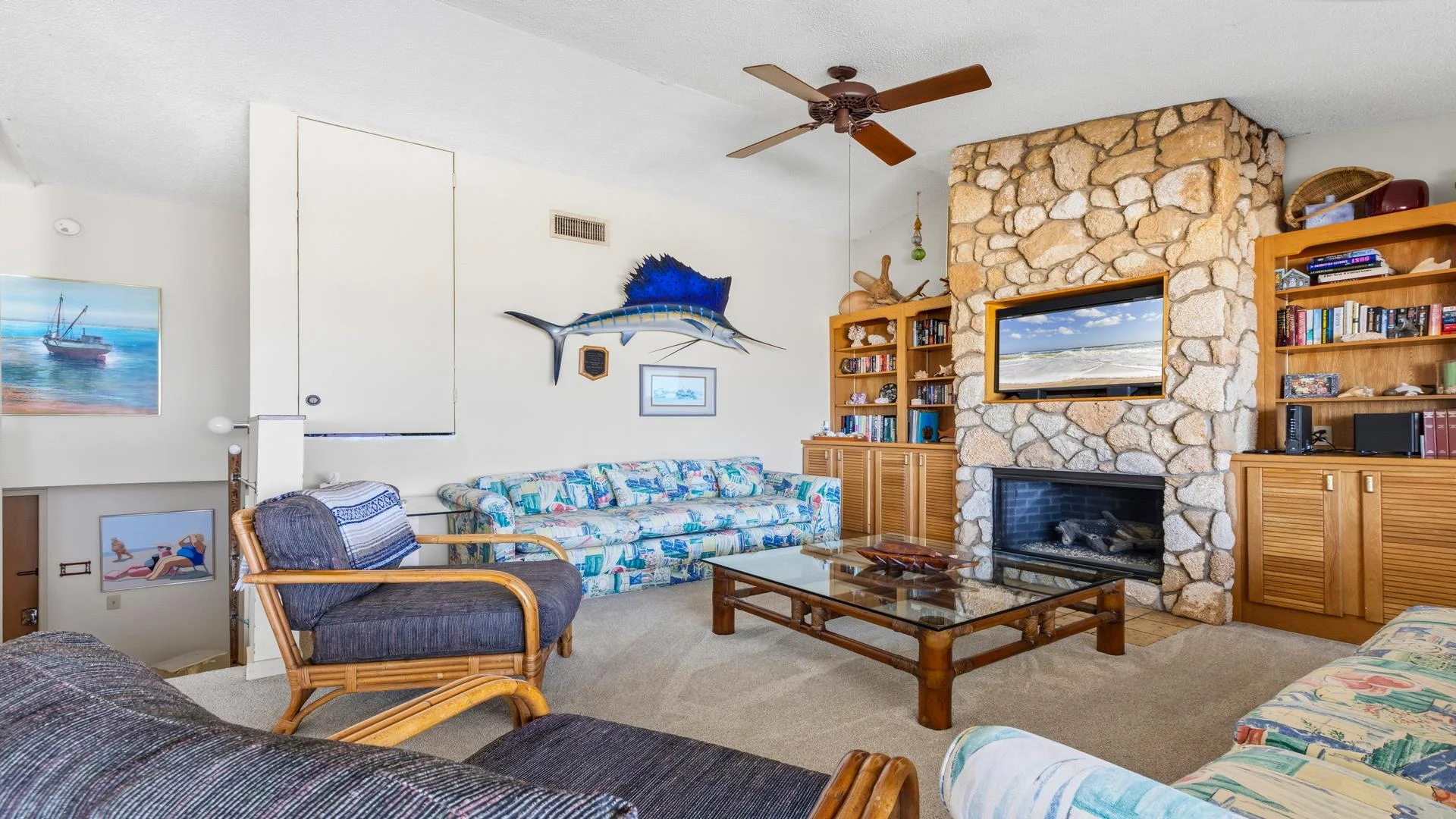 Living room with rattan-framed seating, a glass coffee table, stone fireplace, built-in wood shelves, and mounted fish decor.