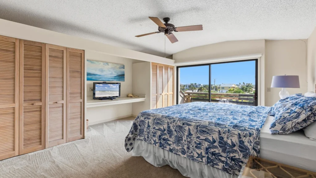 Bedroom with blue sea-themed bedding, beige carpet, louvered closet doors, ceiling fan, and sliding glass door balcony.