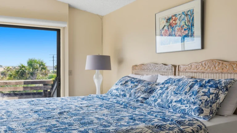 Glass sliding door opens to a wooden balcony railing with palm trees and blue sky beyond the bedroom.